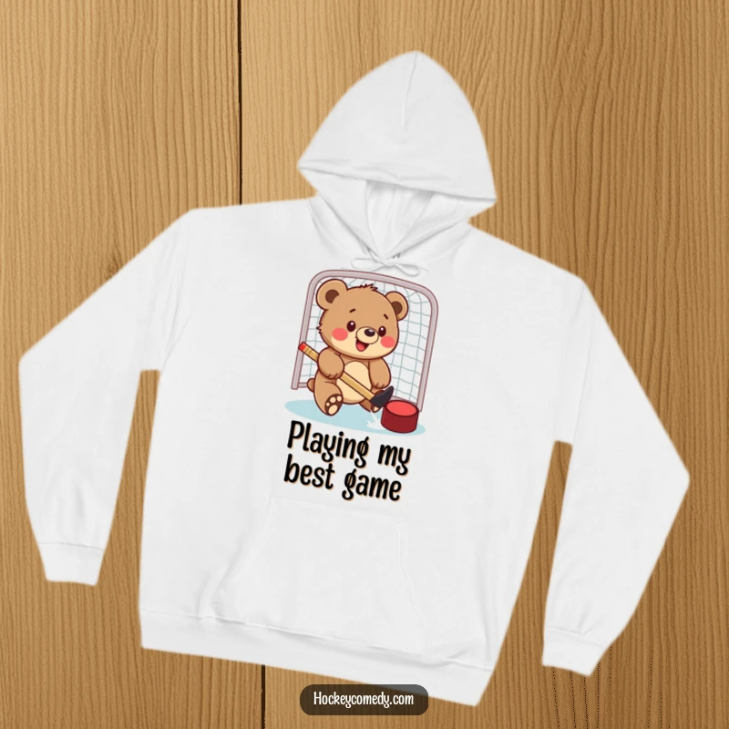 Funny hoodie featuring a happy bear cub playfully hitting a hockey puck into a net, showcasing determination and joy.